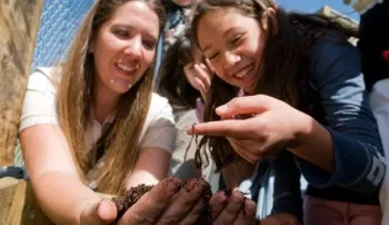 Woman holding handful of worms and compost while teaching a smiling young girl.