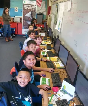 Eight smiling boys sit in a row at computers.