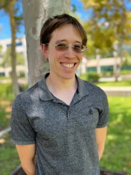 Young man in 30s, smiling at camera while wearing a grey collard shirt and glasses.
