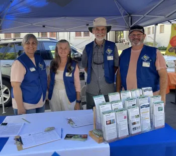 Four smiling people wearing blue vests behind a table with brochures.