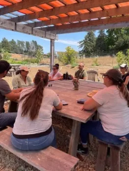 People sit around a table outdoors. Plants grow in the field in the background.