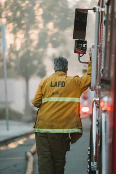 Firefighter leans against fire engine. LAFD printed on back of coat.