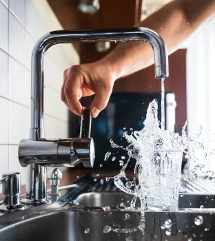 Hand turns on a tap with water splashing out of a glass