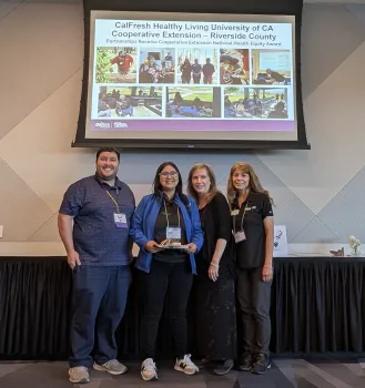 Four people stand in a row under a sign that reads "CalFresh Healthy Living University of CA Cooperative Extension - Riverside County. Partnerships receive Cooperative Extension National Healthy Equity Award" with a collage of photos