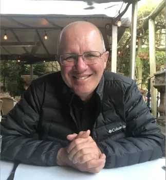 Ken Tate sits at a table with hands folded. He's wearing a brown Columbia jacket