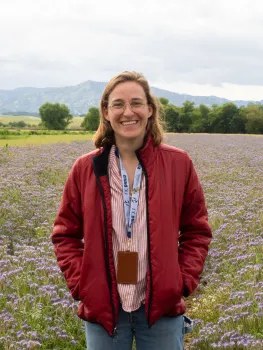 Caroline from waist up wearing a rust-colored jacket and standing in a field of lavender-colored flowers.