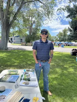 Erik, wearing blue jeans stands on grass beside a table topped with brochures