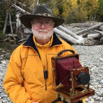 Marshall Johnson wearing a cowboy hat and bright yellow jacket posing with a box camera in a rocky riverbed