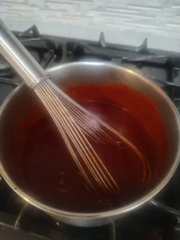 A whisk in red BBQ sauce in a pot on the stove.