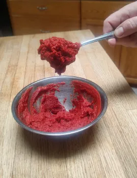 A hand scooping a spoonful of red tomato paste from a metal bowl.