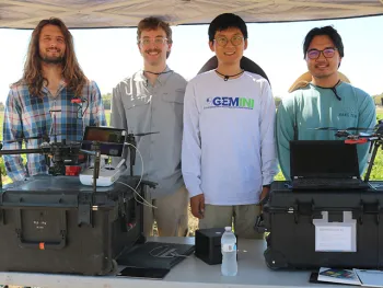 Four young men standing in a row underneath a canopy. In front of them is a table with various instruments