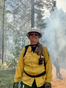 Susie Kocher in helmet and yellow jacket at prescribed burn training as smoke filters through the trees