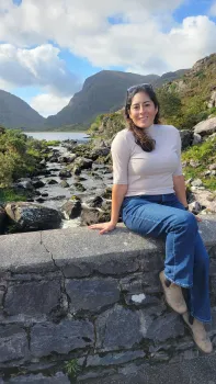 Camila is sitting on a stone wall over a stream bubbling over rocks with blue sky and white clouds overhead