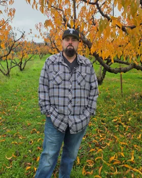 Man in baseball cap and flannel shirt stands with hands in pockets in front of tree with orange leaves in an orchard