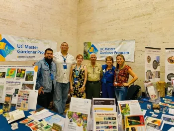 Seven UC Master Gardener volunteers stand behind an outreach table at an indoor event. The table displays educational materials on pollinators, mulching, invasive pests, and tree health, with UC Master Gardener Program banners behind them.