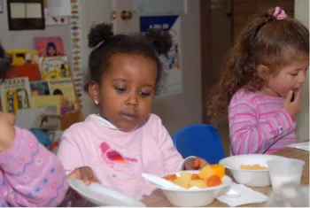 A young girl sitting at a table with other young children grabs a bowl of fruit.