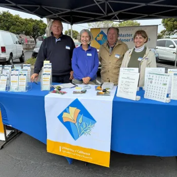 a gorup of 4 volunteers standing in a uc master gardenrr booth at a farmers market