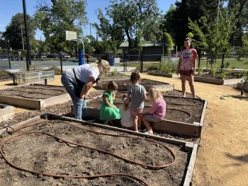 A UC Master Gardener volunteer guides four young students gathered around a raised garden bed, exploring soil during a hands-on lesson. Another adult observes nearby. The school garden includes multiple raised beds with irrigation lines, surrounded by a fenced outdoor space with trees and a basketball court in the background.