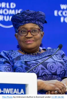 Ngozi Okonjo-Iweala sitting at the podium of the world economic forum in 2021. She is wearing a blu dress with swirl patterns and a blue head covering with a smile on her face. 