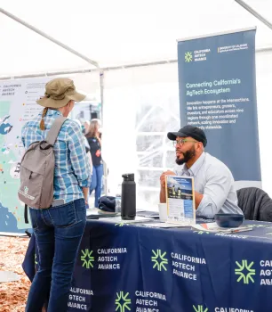 Hanif, seated behind the California AgTech Alliance table, talks with a woman wearing a backpack standing on the other side of the table.