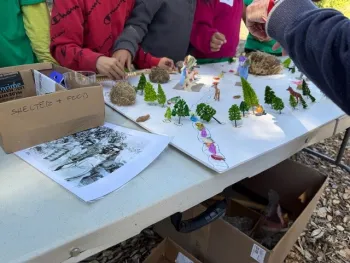 Students gather around a table building a miniature Wappo village model using small trees, animals, and natural materials to represent food, shelter, and habitat. Hands-on materials, drawings, and labeled supplies support learning about Indigenous culture and ecosystems during an outdoor garden lesson.