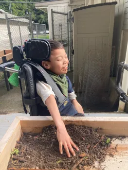 Young boy seated and strapped into a wheelchair seated adjacent to an elevated garden box, touching dirt with his left hand and smiling with joy