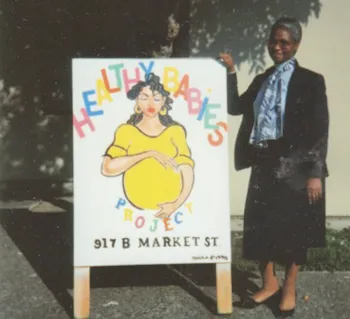 Mary Blackburn stands next to a sign that reads: Healthy babies project. 917 B Market St. with a drawing of a dark-haired woman cradling her pregnant belly