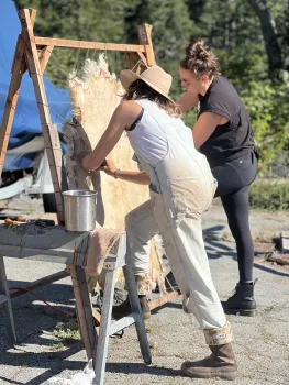 A person working on a sheep hide stretched across a rack, another person helps to support the hide.