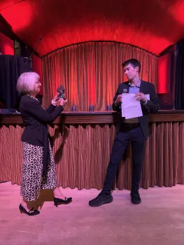 A woman holds a sculpted award toward Daniel, who is holding sheets of paper. Red lights illuminated curtains behind them