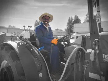 Man wearing a beige hat, orange gloves and blue coveralls sits in the driver's seat of a spray rig. Most of the photo is black and white, except for the man.