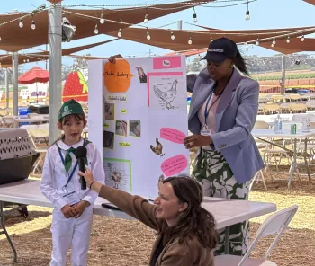 Girl in 4-H uniform speaks into mic held by an adult woman while another woman holds up the chicken project poster on a table
