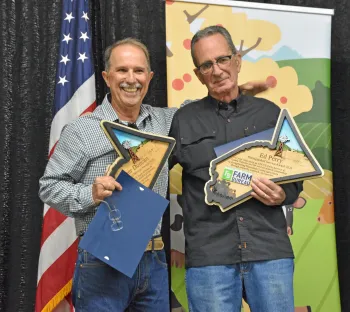 Two smiling men stand in front of a U.S. flag holding California-shaped plaques