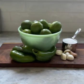 a bowl of limes, three jalapenos, peeled cloves of garlic and a cup of salt on a cutting board