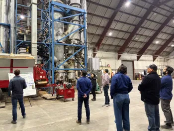 a group of people in a large industrial space look at a system of large pipes in a scaffolding about 50 feet high while someone explains the system