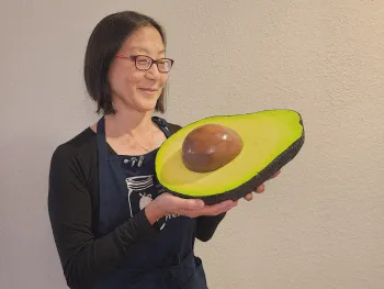 Marina holding a large wooden half-avocado smiles at the camera
