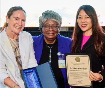 Three women, one holding a blue box containing a lucite trophy, another holding a certificate.