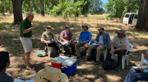 Man in polo and shorts and hat at left speaks with a group of students under shade trees during a farm tour