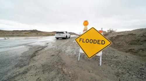 A road sign that reads "Flooded" in front of a car that is on a flooded street