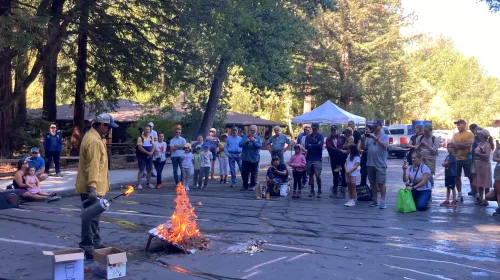 A crowd of kids and adults watch a fire expert light a fire on an inclined board 