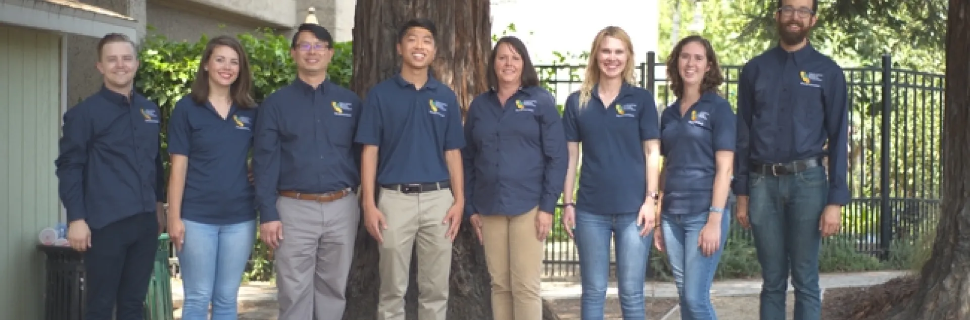 8 people in dark blue shirts standing in a row under a large tree