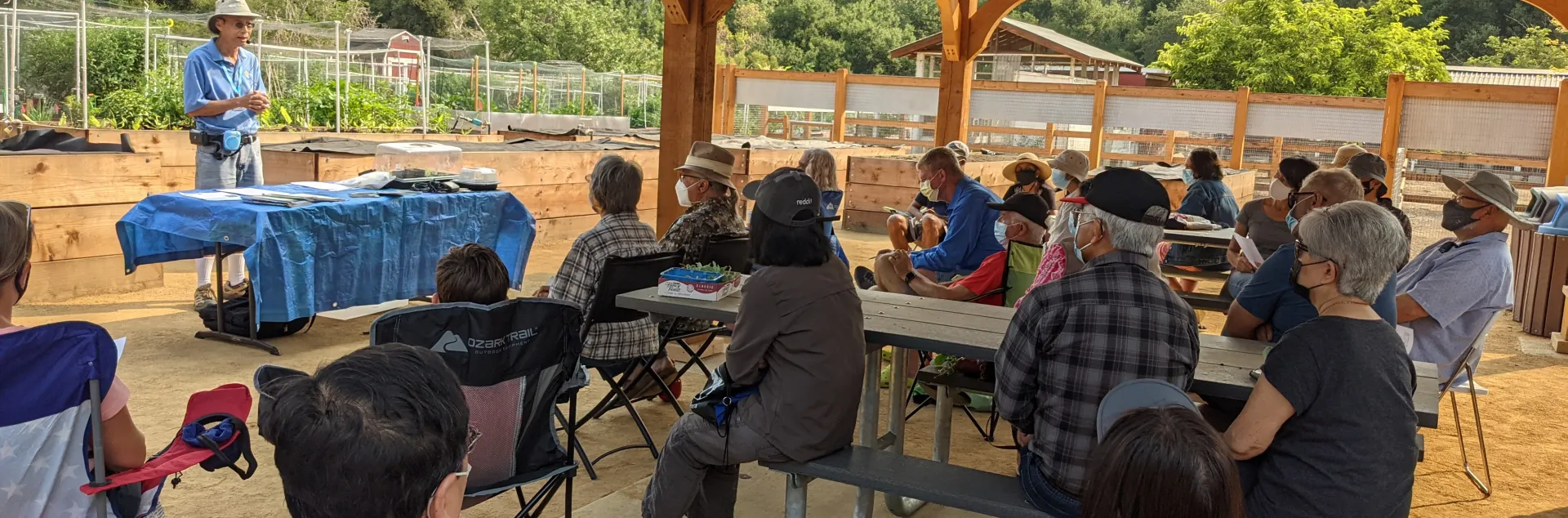 A man presents a sustainable gardening class to a seated audience under a wooden pavilion at McClellan Ranch Preserve, surrounded by lush gardens.
