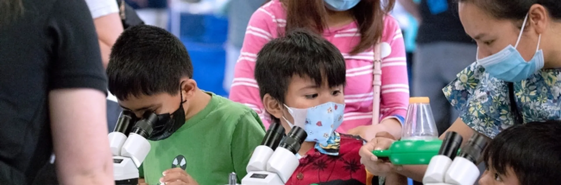 Three dark-haired kids assisted by two women use microscopes.