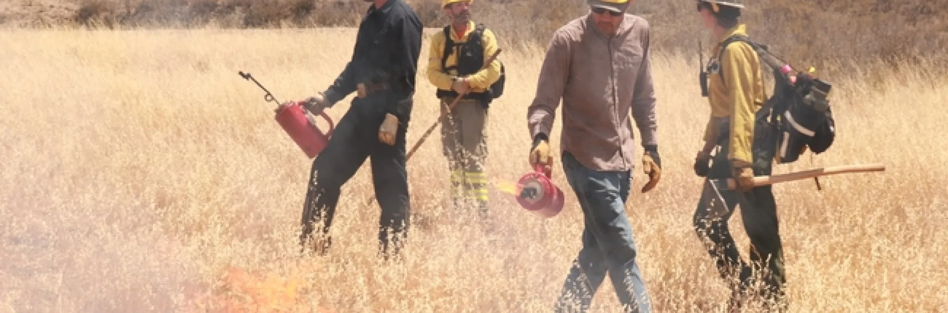 Three people wearing hard hats and carrying drip torches.
