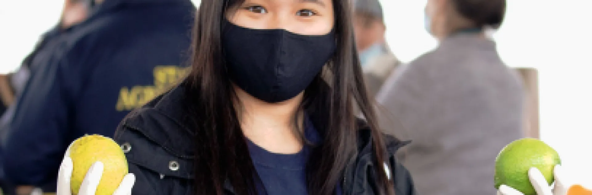 A young woman wearing a facemask holds a piece of citrus in each hand.
