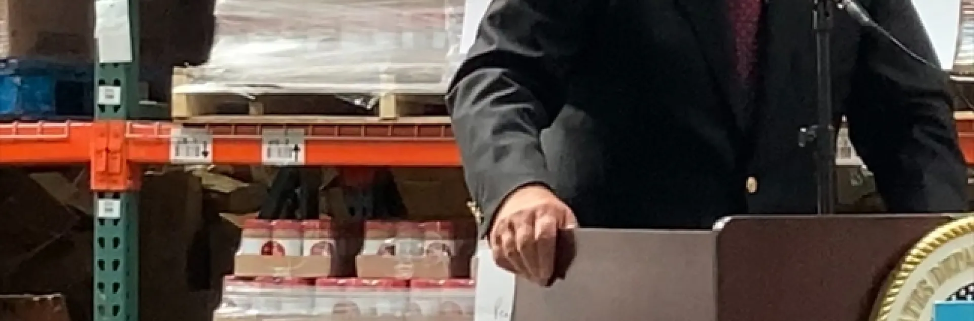 Vilsack stands at lectern with pallets of canned goods on shelves behind him.