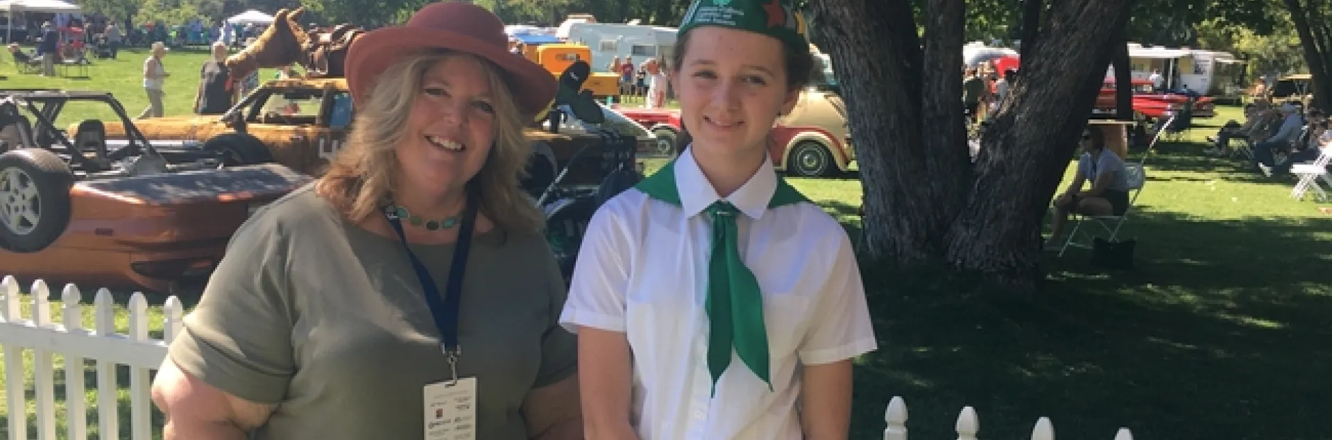 June, wearing a hat, stands under a leafy tree beside Lily, who is wearing a 4-H uniform of green and white. Classic cars are in the background.