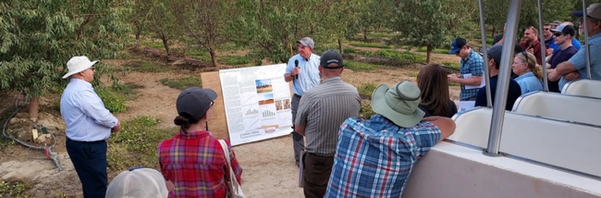 People stand in an almond orchard listening to Brent Holtz, who is holding a mic.