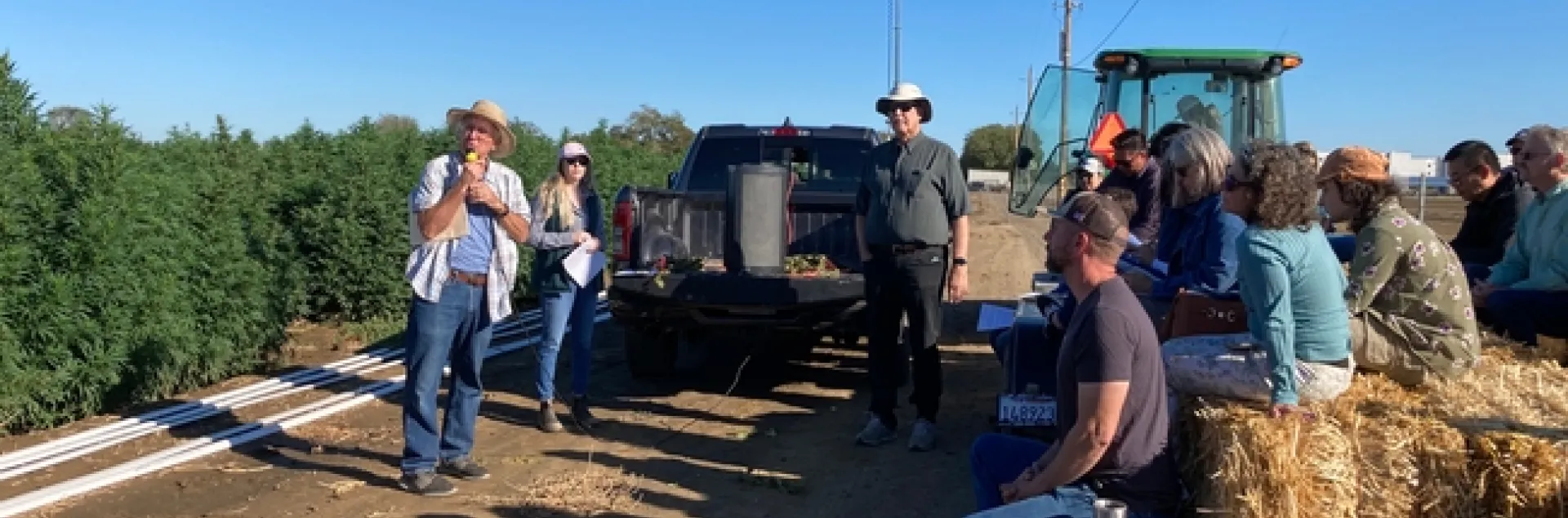 Standing beside a field of hemp, Bob Hutmacher speaks into a mic as growers and others sit on hay bales listening.