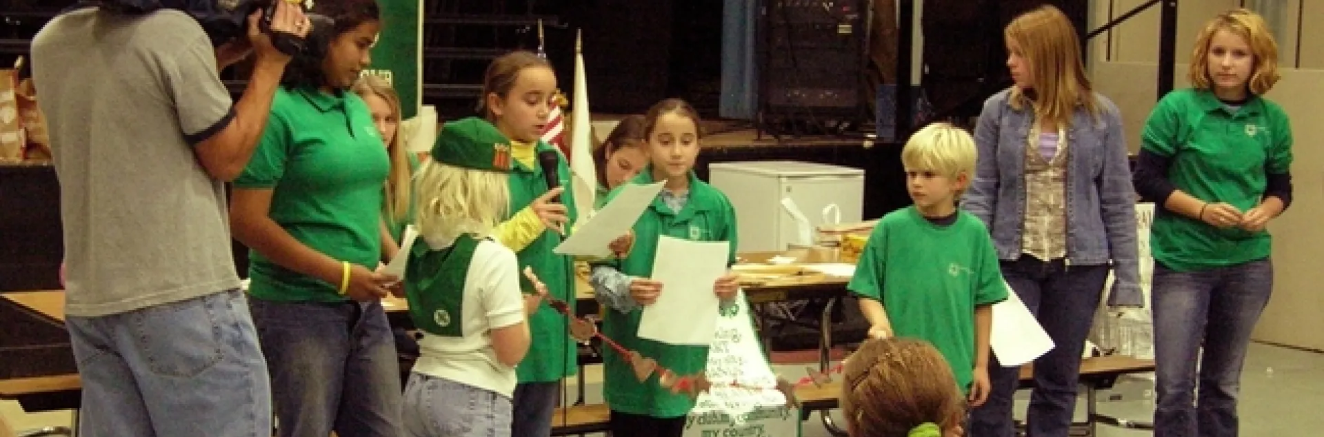 Kids in green shirts read from pieces of paper as someone shoots video.