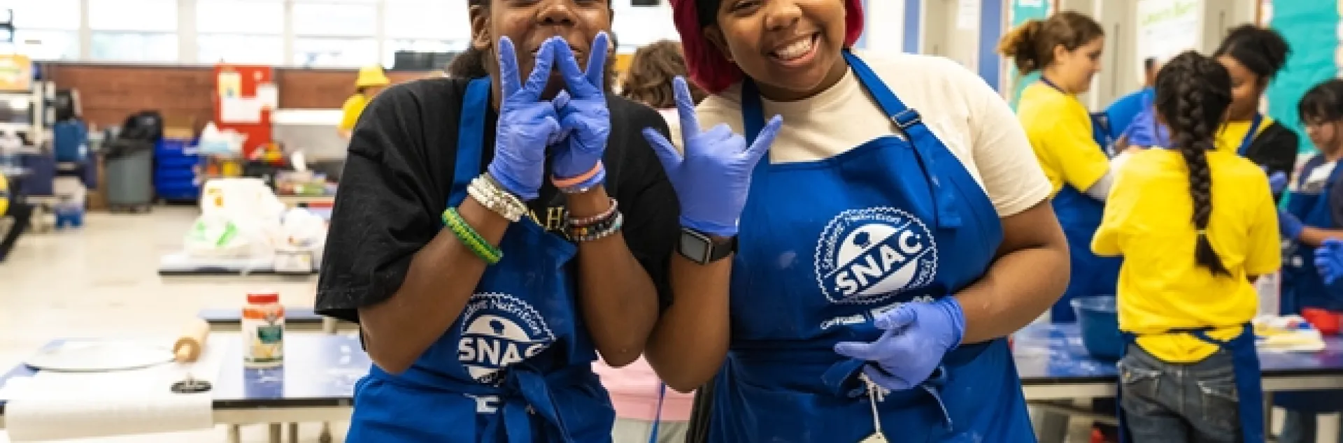Two girls smiling in a school cafeteria with blue aprons and food prep gloves on.